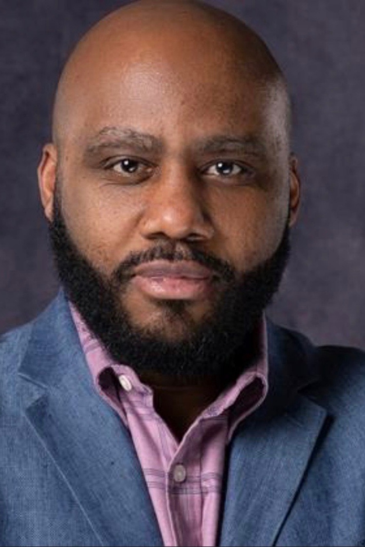 A headshot of Adrian, wearing a blue jacket and a pink shirt.