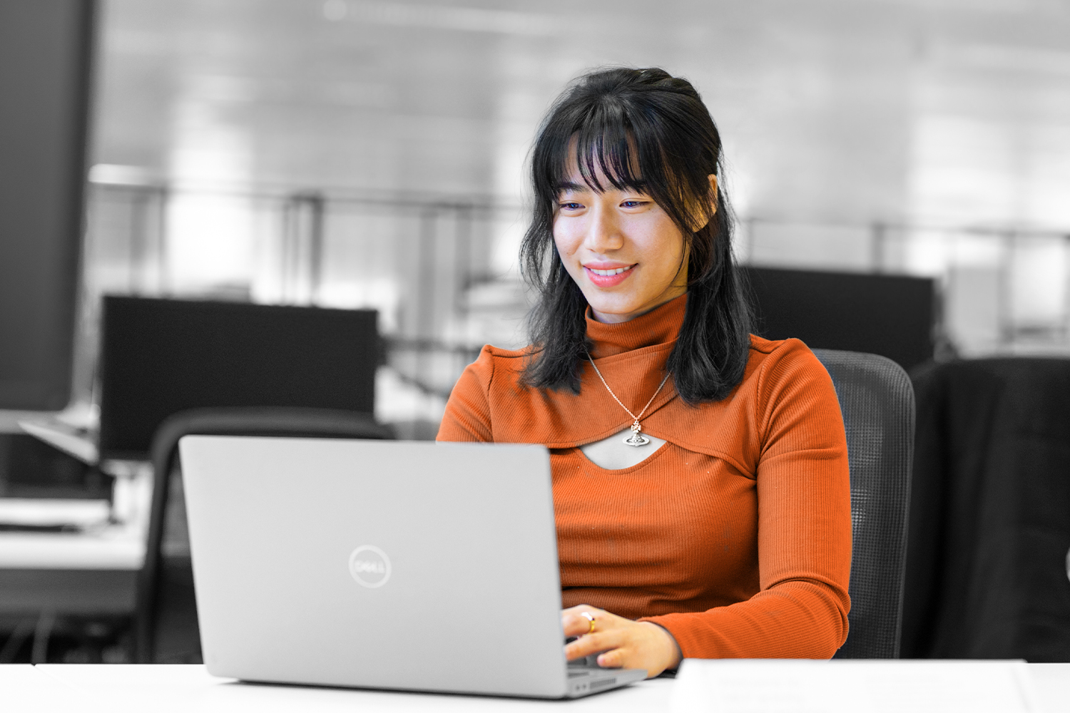 Black and White image, where only clothes and skin tone are in colour. A young woman in an orange top works at a laptop. 